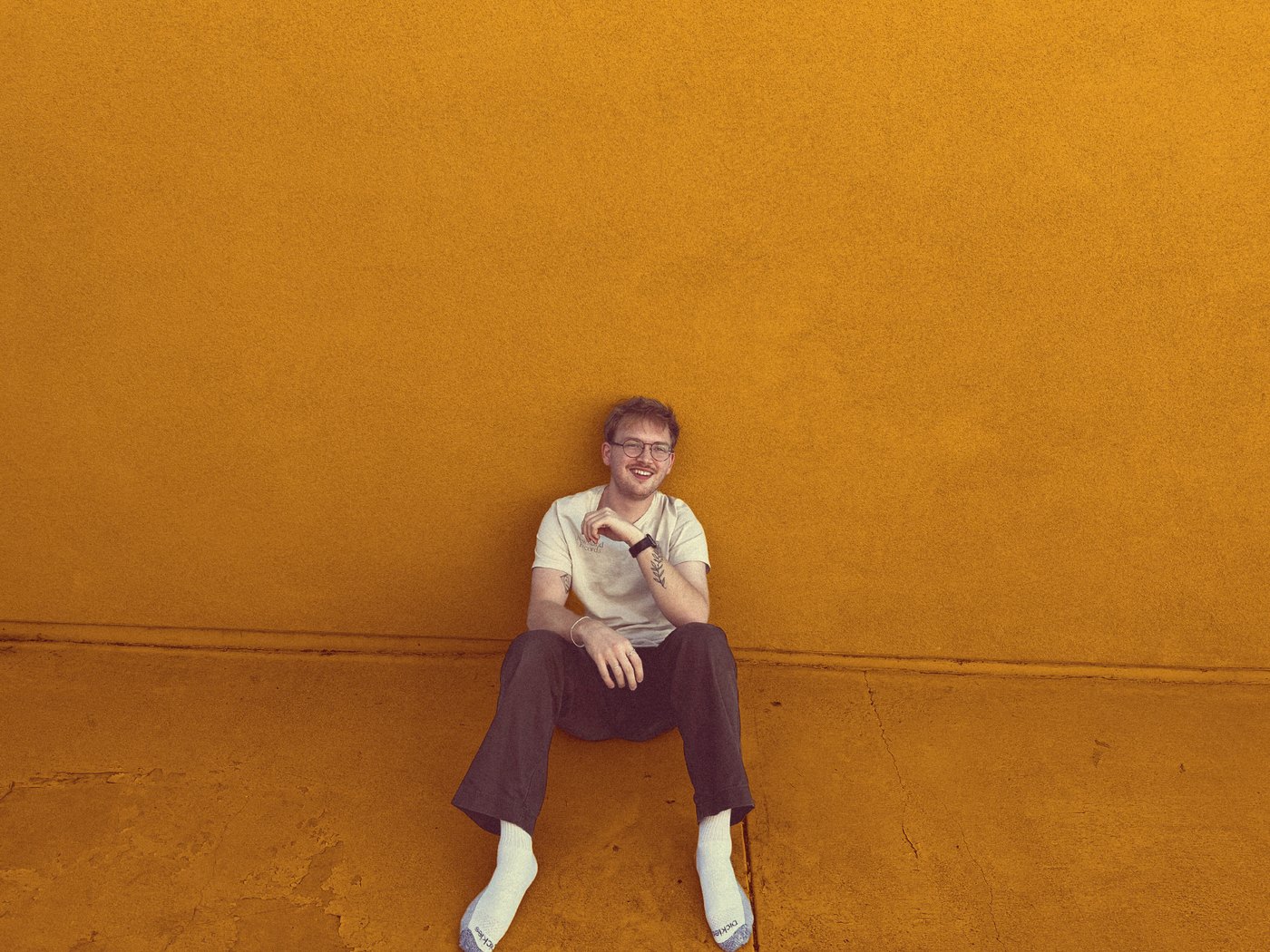Geepers! sitting against a mustard-yellow wall in a white tee, dark pants and white socks, smiling